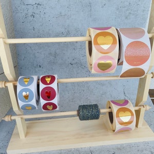 Könnte beinhalten: Ein Holzständer mit vier Rollen Aufkleber. Die Aufkleber sind rund und haben ein Herzdesign. Die Aufkleber sind in verschiedenen Farben, darunter Gold, Rosa, Rot und Blau.