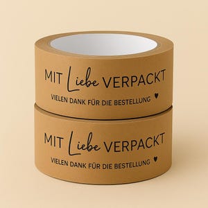以下が含まれることがあります： 茶色の梱包用テープが2巻、重ねられています。「MIT Liebe VERPACKT」と「VIELEN DANK F&Uuml;R DIE BESTELLUNG」という文字が黒い筆記体で印刷されています。小さなハートのシンボルも付いています。