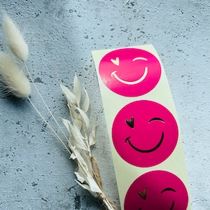 Könnte beinhalten: Drei runde rosa Aufkleber mit weißen, zwinkernden Smiley-Gesichtern und Herzen als Augen. Die Aufkleber befinden sich auf einem weißen Blatt.