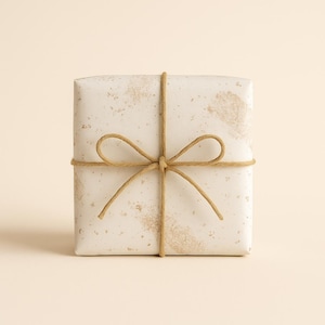 Pode incluir: Uma caixa de presente quadrada embrulhada em papel branco sujo com um padrão salpicado. A caixa é amarrada com barbante natural, formando um laço na parte superior. O fundo é de cor bege claro sólido.