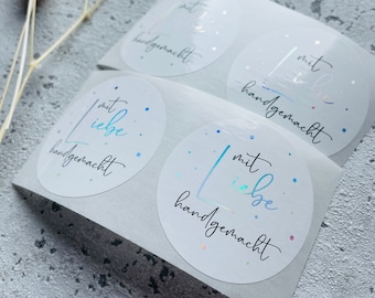 May include: Several white, round stickers with the words "mit Liebe handgemacht" in a handwritten font. The stickers have a holographic sheen and are scattered with small, colorful dots. The background is a light gray surface.