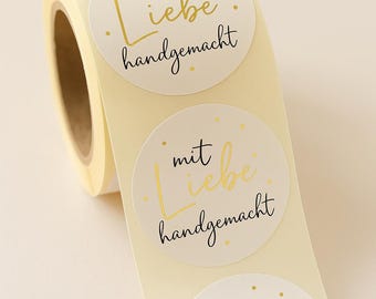 Puede incluir: Un rollo de pegatinas redondas de color crema con texto dorado y negro. Cada pegatina dice "mit Liebe handgemacht" en un estilo manuscrito. Las pegatinas están diseñadas para artículos hechos a mano.