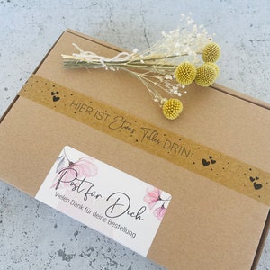 Pu&ograve; includere: Una scatola di cartone marrone con un adesivo che dice "Post f&uuml;r Dich" e "Vielen Dank f&uuml;r deine Bestellung". La scatola &egrave; decorata con un piccolo mazzo di fiori secchi bianchi e gialli e un nastro di carta marrone con cuori neri.