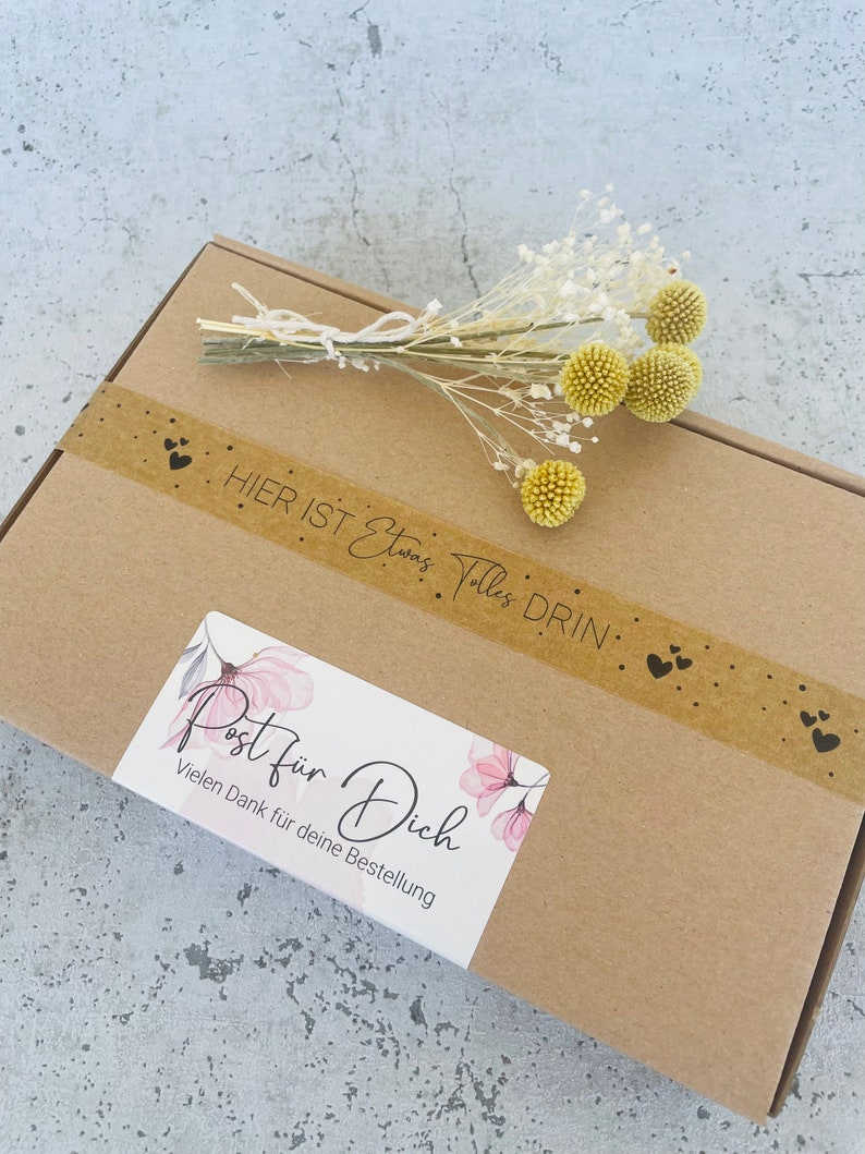 Pu&ograve; includere: Una scatola di cartone marrone con un adesivo che dice "Post f&uuml;r Dich" e "Vielen Dank f&uuml;r deine Bestellung". La scatola &egrave; decorata con un piccolo mazzo di fiori secchi bianchi e gialli e un nastro di carta marrone con cuori neri.