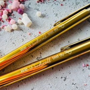 Peut inclure: Deux stylos dorés avec l'inscription "Wie wundervoll bist du" (Comme tu es merveilleux) en lettres blanches. Chaque stylo a un petit symbole de cœur à la fin de l'inscription.