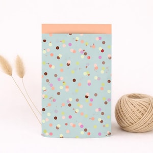 Op de afbeelding: Een rechthoekig notitieblok met een lichtblauwe achtergrond en kleurrijke stippen. De bovenrand is perzikkleurig. Een bol touw en twee gedroogde bloemstelen liggen naast het notitieblok.