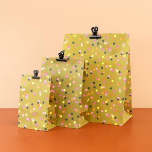 Könnte beinhalten: Drei Geschenkpapierbeutel in verschiedenen Größen, verziert mit bunten Konfetti-Punkten auf einem senfgelben Hintergrund. Jeder Beutel wird mit einer schwarzen Klammer oben verschlossen, was einen festlichen und verspielten Look erzeugt.