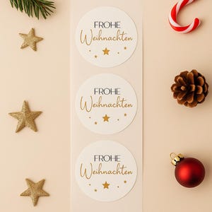 Könnte beinhalten: Drei weiße runde Aufkleber mit dem Text "FROHE Weihnachten" in goldener Schrift und goldenen Sternen. Die Aufkleber befinden sich auf einem Streifen, umgeben von goldenen Sternen, einem Tannenzweig, einer Zuckerstange und einem Tannenzapfen.