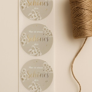May include: Three round stickers with the text "Hier ist etwas Schönes drin" in gold script and a floral design. A roll of brown twine is visible on the right side of the image. The stickers are on a white backing.