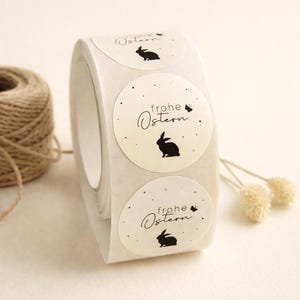 Może przedstawiać: Rolka okrągłych, kremowych naklejek z czarną sylwetką królika i napisem "frohe Ostern" w eleganckiej czcionce. Naklejki mają małe czarne kropki i motyla. W tle rolka sznurka.