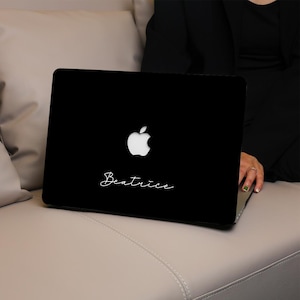 May include: Black laptop with a white Apple logo and the name "Beatrice" in cursive script. The laptop is resting on a gray surface, with a person wearing a black top in the background.