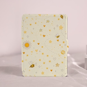 以下が含まれることがあります： ミツバチ、星、ハート、花をあしらった、気まぐれなデザインのライトイエローのタブレットケース。ケースはマグネット開閉式で、柔らかい模様の入った外装です。デザインには大きなデイジーと点線が含まれています。