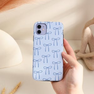 Pode incluir: Uma capa de telefone azul claro com um padrão repetido de laços azul marinho. A capa tem um acabamento brilhante e foi projetada para um smartphone. A capa do telefone é segurada por uma pessoa.