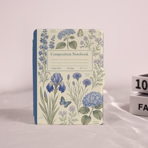 May include: A composition notebook with a cream-colored cover featuring blue botanical illustrations of irises, hydrangeas, and butterflies. The center label reads Composition Notebook, College Ruled, 120 Pages, 8.5 x 11 inches, with a blue spine.