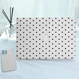 Puede incluir: Funda blanca para portátil y funda para teléfono con un patrón de lunares negros. La funda del portátil tiene un recorte para el logotipo de Apple. La funda del teléfono está al lado de la funda del portátil.