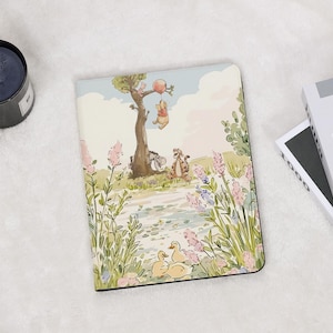 Puede incluir: Un cuaderno ilustrado a color con Winnie the Pooh y sus amigos. La portada representa una escena con Pooh colgado de un árbol con un globo, y otros personajes en un entorno exterior caprichoso con patos y flores.