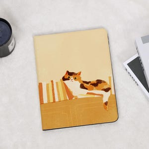 May include: A rectangular tablet case with a light yellow background featuring a printed illustration of a calico cat resting on a shelf next to a row of books. The cat has white, orange, and dark brown patches and is looking forward.