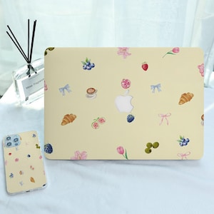 Puede incluir: Una funda para portátil y una funda para teléfono de color amarillo claro con un patrón repetido de ilustraciones en acuarela. El diseño incluye cruasanes, tazas de café, bayas, flores y lazos. La funda del portátil tiene un logotipo blanco de Apple.