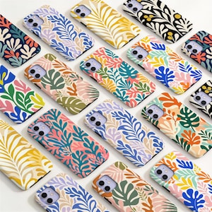 May include: A collection of smartphone cases featuring various colorful botanical and leaf patterns. Each case displays a unique arrangement of stylized foliage in shades of blue, green, yellow, orange, pink, and cream, with cutouts for camera lenses.