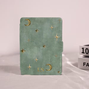 May include: A sage green e-reader cover featuring a textured surface with gold foil celestial motifs, including multiple four-pointed stars and two crescent moons. The letters ES are printed in gold near the bottom center of the cover.