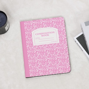 Peut inclure: Un cahier de composition rose avec un motif moucheté blanc et une étiquette blanche qui dit "COMPOSITION BOOK". Le cahier est ouvert, révélant le dos rose et les bords noirs. Le cahier est sur une surface blanche.