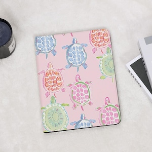 May include: A pink tablet case featuring a pattern of colorful watercolor sea turtles. The turtles are in shades of blue, orange, green, and pink. The case has a black border and is on a white surface.