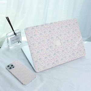 Könnte beinhalten: Ein Laptop- und Handyhüllen-Set mit einem sich wiederholenden Muster aus rosa und blauen Schleifen auf hellem Hintergrund. Der Laptop ist geöffnet und das Telefon liegt daneben. Ein Glasdiffusor mit Schilf befindet sich im Hintergrund.