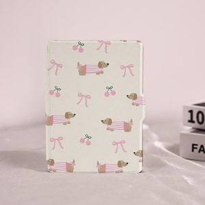 Könnte beinhalten: Eine cremefarbene Tablet-Hülle mit einem sich wiederholenden Muster aus Dackeln in rosa gestreiften Hemden, rosa Schleifen und rosa Kirschen. Die Hülle hat einen Magnetverschluss.