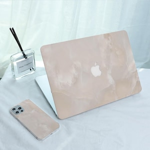Può includere: Un laptop e uno smartphone con cover abbinate con motivo marmorizzato beige. Il laptop ha un logo Apple bianco. Nell'immagine è presente anche una bottiglia di vetro trasparente con bastoncini neri e la scritta "Mose Fragrance".