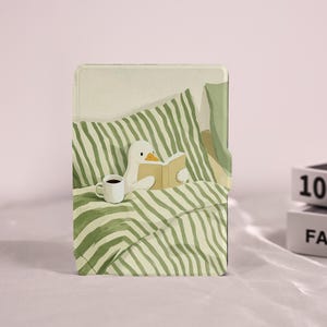 Puede incluir: Una funda rectangular para lector electrónico con una ilustración impresa de un pato blanco sentado en una cama con ropa de cama a rayas verdes y blancas. El pato sostiene un libro abierto y una taza blanca descansa sobre la cama.