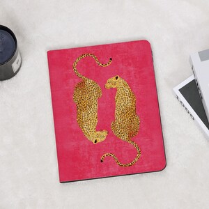 May include: A pink notebook featuring two leopard illustrations with yellow and brown spots. The leopards are positioned facing each other. The notebook is rectangular and has a black border.