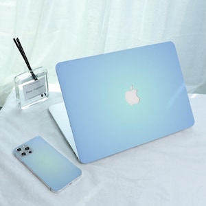 Könnte beinhalten: Ein hellblaues Laptop mit weißem Apple-Logo ist auf einer weißen Oberfläche geöffnet. Ein passendes hellblaues Smartphone und ein klarer Glasdiffusor mit schwarzen Stäbchen sind ebenfalls vorhanden. Der Diffusor trägt den Text "Mose Fragrance".