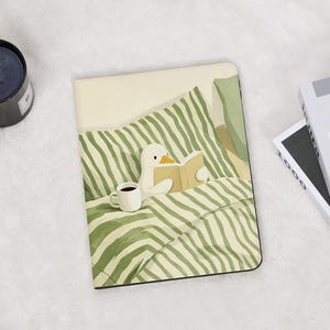 May include: A rectangular e-reader case featuring an illustration of a white duck sitting in a bed with green and white striped bedding. The duck is holding an open book, and a white mug filled with a dark beverage sits on the striped blanket.