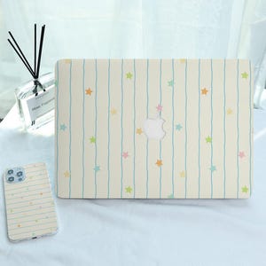 Op de afbeelding: Een crèmekleurige laptophoes en telefoonhoesje met een patroon van verticale lichtblauwe strepen en kleurrijke sterren. De laptophoes heeft een uitsparing voor het Apple-logo. De telefoonhoes zit links.