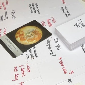 Puede incluir: Una colecci&oacute;n de tarjetas blancas con frases escritas a mano y una pila de tarjetas con "I LOVE YOU!" escrito en ellas. Tambi&eacute;n hay una sola tarjeta con una ilustraci&oacute;n de luna llena. Las tarjetas est&aacute;n esparcidas sobre una superficie blanca.