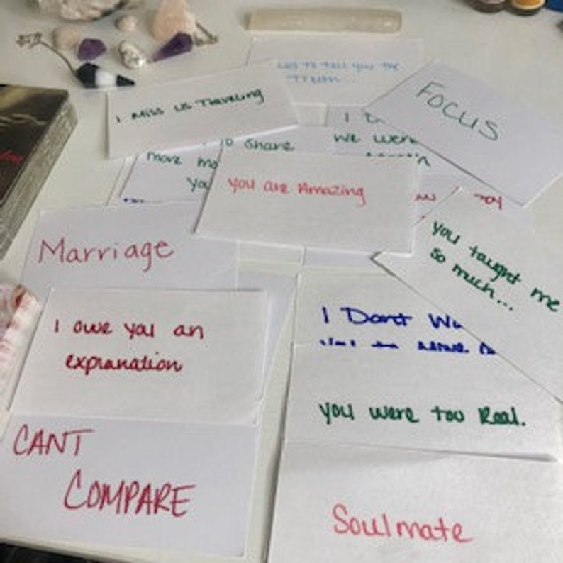 Puede incluir: Una colecci&oacute;n de tarjetas blancas con mensajes escritos a mano en varios colores, incluyendo "I MISS US TRAVELING", "You are Amazing" y "Soulmate". Las tarjetas est&aacute;n esparcidas sobre una superficie blanca, con una baraja de cartas y cristales visibles.
