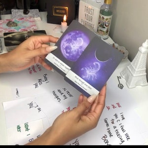 Puede incluir: Se muestran dos cartas de tarot con dise&ntilde;os celestiales. Una carta presenta una luna llena p&uacute;rpura y el texto "It's time to release anyone". La otra carta muestra una luna creciente y un escorpi&oacute;n con el texto "Work through your fears".