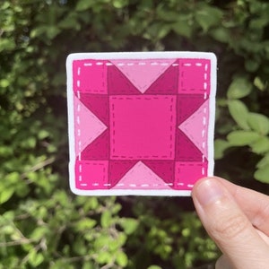 May include: Pink and white quilt block sticker with a star design. The sticker has a white border and a stitched look.