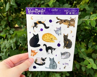 May include: A sticker sheet featuring a variety of cats in different poses. The sheet is white with a purple border and includes the text "Kellyanne Makes Art + cats + sticker sheet + @kellyannemakesart".