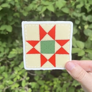 May include: A square sticker featuring a quilt block design. The sticker has a cream-colored background with a red star pattern and a green center square. The edges are outlined with a dotted line.