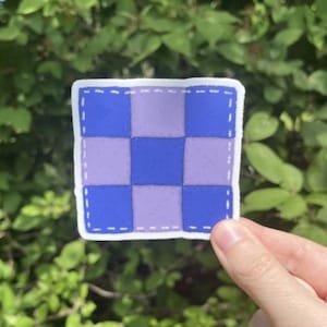 May include: A purple and blue checkered sticker with a white border and a stitched design.