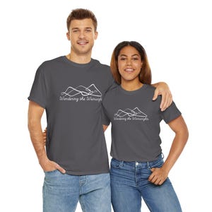 May include: Two people wearing charcoal grey t-shirts with the text "Wandering the Wainwrights" and a mountain graphic in white. The shirts are paired with blue jeans. The image is taken from the waist up.