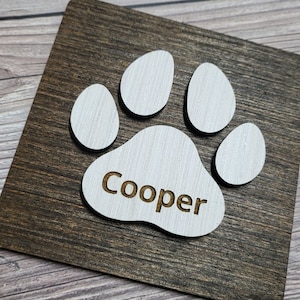 May include: A wooden sign with a white paw print and the name "Cooper" engraved in the centre. The sign has a dark brown wood grain.