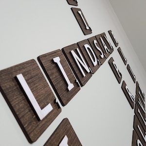 May include: A wall-mounted display of dark brown wooden square tiles featuring white raised letters and small numerical values in the bottom right corner, arranged in a crossword-style grid spelling out names including LINDSAY and other words.