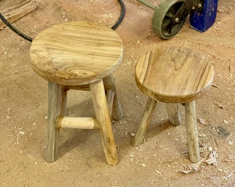May include: Two round wooden stools with natural grain patterns and light brown tones. The larger stool features three legs connected by horizontal support bars, while the smaller stool has three simple legs without additional support structures.