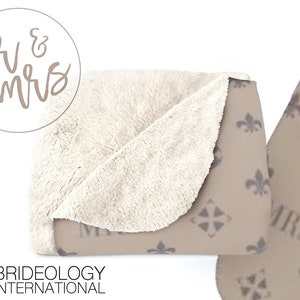Puede incluir: Una manta suave y mullida con un lado de felpa beige y un lado impreso con un patr&oacute;n repetido de flores de lis y las palabras "Mr. & Mrs."