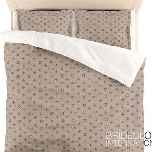 Puede incluir: Una funda n&oacute;rdica de color marr&oacute;n claro con un patr&oacute;n repetido de una flor de lis y las palabras "MR" y "MRS". La funda n&oacute;rdica est&aacute; en una cama con dos fundas de almohada a juego. El texto "BRIDEOLOGY INTERNATIONAL" es visible en la esquina inferior derecha de la imagen.