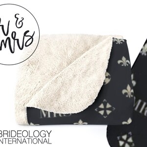 May include: A black and white throw blanket with a repeating pattern of fleur-de-lis and a stylized cross. The blanket has a white sherpa lining and the words "Mr. & Mrs." are printed on the front.