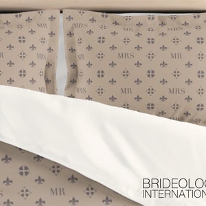 Puede incluir: Dos fundas de almohada de color beige con un patr&oacute;n repetido de una flor de lis y las palabras "MR" y "MRS" en color gris oscuro. Las fundas de almohada est&aacute;n sobre una cama blanca con las palabras "BRIDEOLOGY INTERNATIONAL" impresas en negro en la parte inferior.