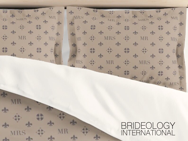 Puede incluir: Dos fundas de almohada de color beige con un patr&oacute;n repetido de una flor de lis y las palabras "MR" y "MRS" en color gris oscuro. Las fundas de almohada est&aacute;n sobre una cama blanca con las palabras "BRIDEOLOGY INTERNATIONAL" impresas en negro en la parte inferior.